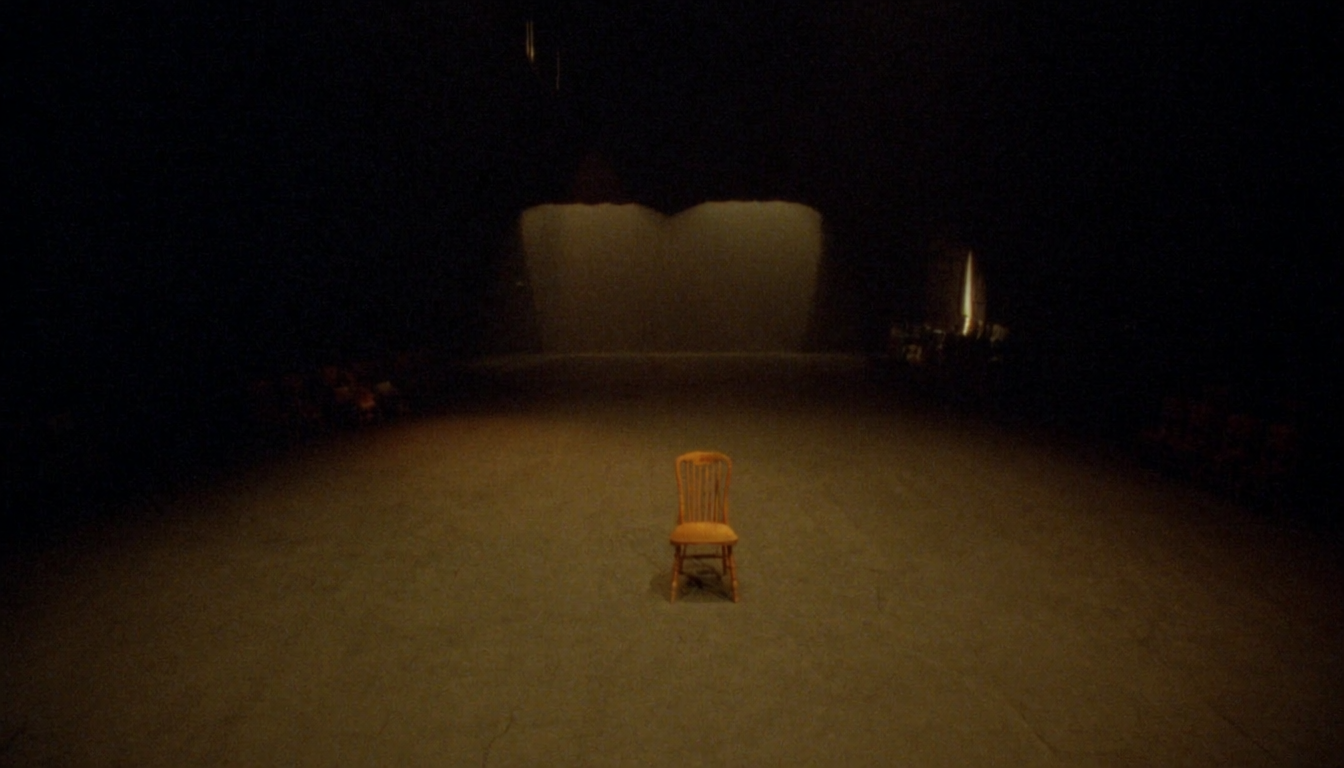Empty rehearsal room with a single wooden chair center frame, warm amber stage lighting and dark wings receding into shadow
